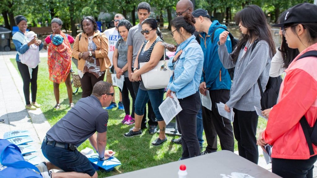 A man kneels on the grass demonstrating CPR to a group of attentive people. They are outdoors, surrounded by trees, holding pamphlets. A table with a water bottle is in the foreground.