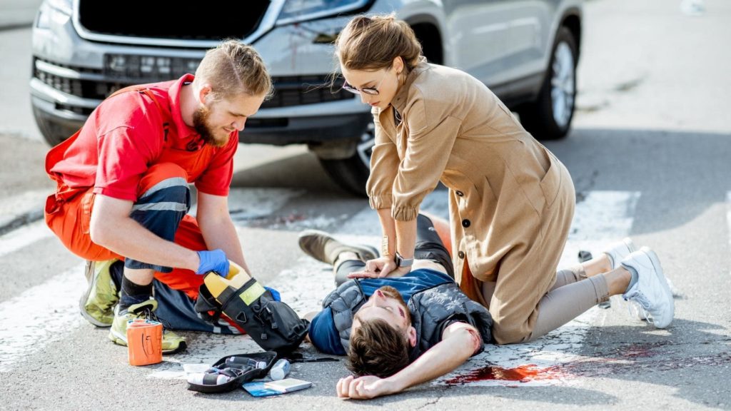 A paramedic and a woman attend to an injured person lying on a crosswalk near a car. The paramedic is checking the injured person's arm, while the woman is supporting their head. Medical supplies are scattered on the ground.