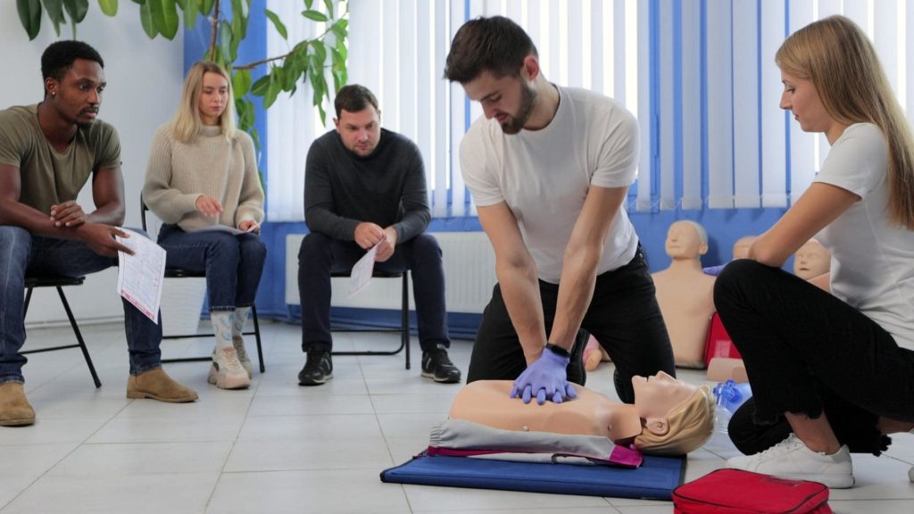 A group of people in a classroom setting are practicing CPR on a mannequin. A man in a white shirt and gloves demonstrates chest compressions, while others watch, holding instruction sheets. A red bag is on the floor nearby.