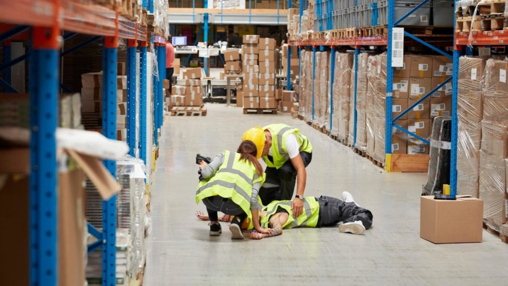 Two workers in high-visibility vests and helmets attend to a person lying on the floor in a warehouse aisle. Shelves loaded with boxes line both sides, and a few cardboard boxes are on the ground nearby.