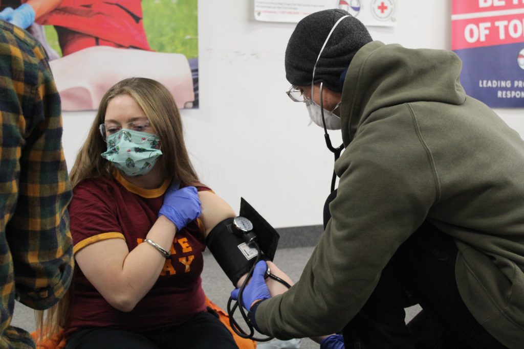 A person wearing a mask and gloves checks the blood pressure of a woman seated beside them, who is also masked. They are in an indoor setting with a poster in the background. Both appear focused on the activity.
