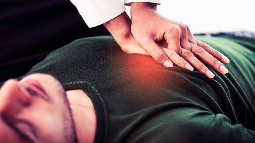A person is lying on their back wearing a green shirt, while another person's hands are performing CPR on their chest. The scene suggests a medical emergency or life-saving procedure.