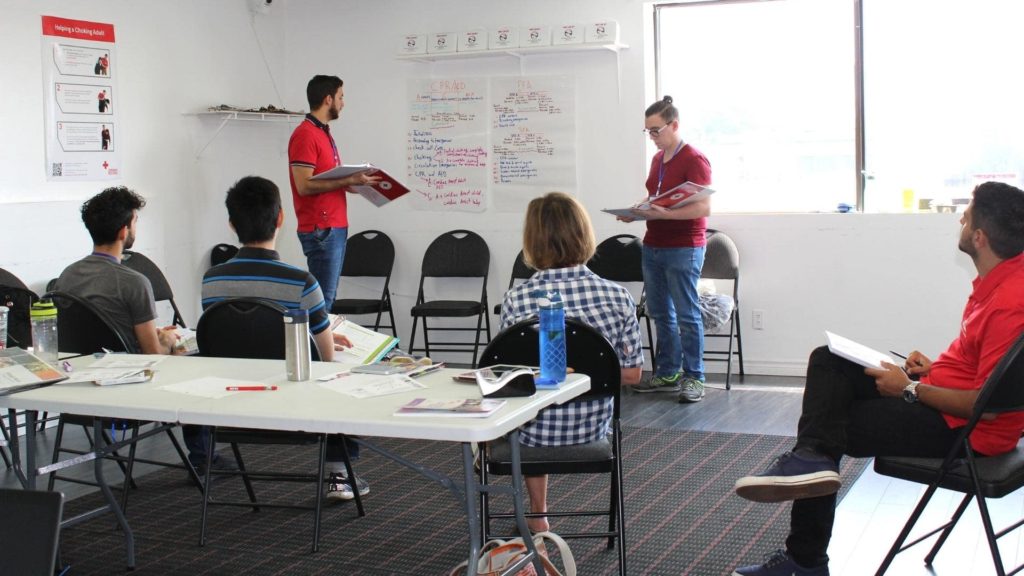 A group of people in a classroom setting, with two presenters standing near a whiteboard covered in notes. The audience is seated at a table with papers and water bottles. A large window brings in natural light.