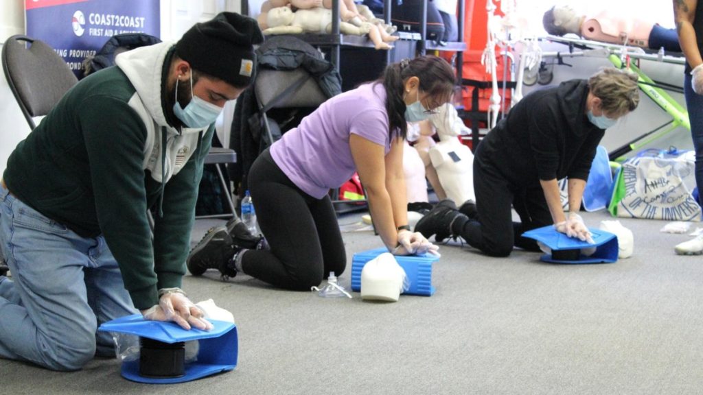 Three people kneel on the floor practicing CPR on training dummies. They wear masks and gloves. Various CPR equipment is visible in the background.