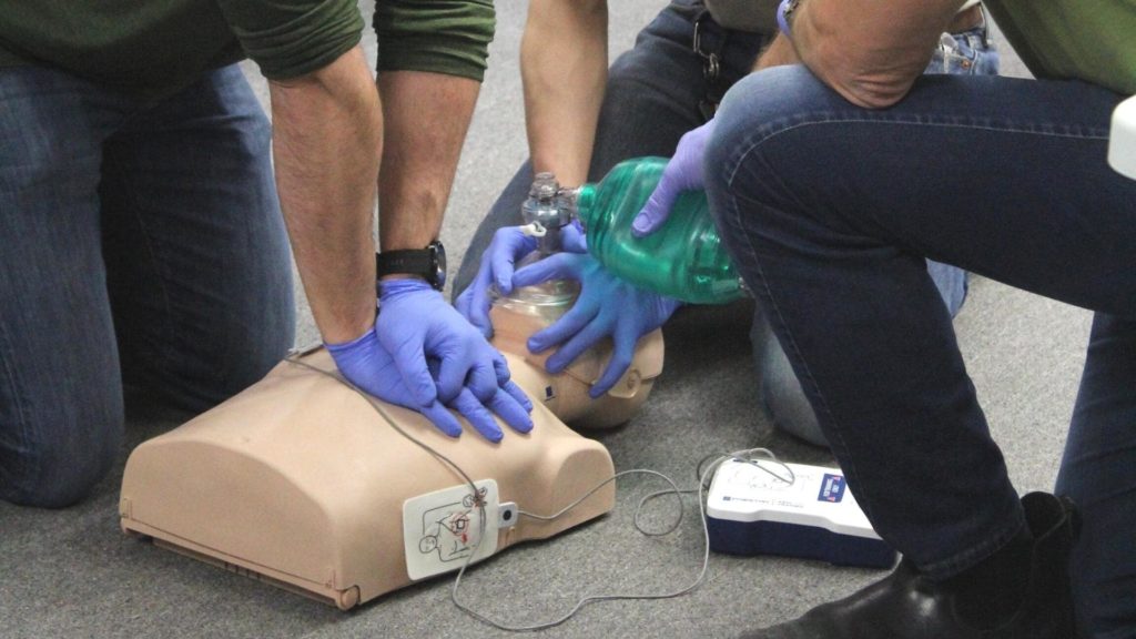 Three individuals practice CPR on a mannequin. Two compress the chest, and one uses a bag mask for ventilation. An automated external defibrillator (AED) is positioned nearby. All are wearing blue gloves.