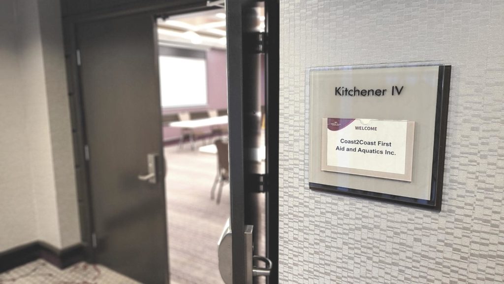 Open door to conference room labeled “Kitchener IV” with a sign for Coast2Coast First Aid and Aquatics Inc. Inside, tables and chairs are arranged, with a projector screen in the background.