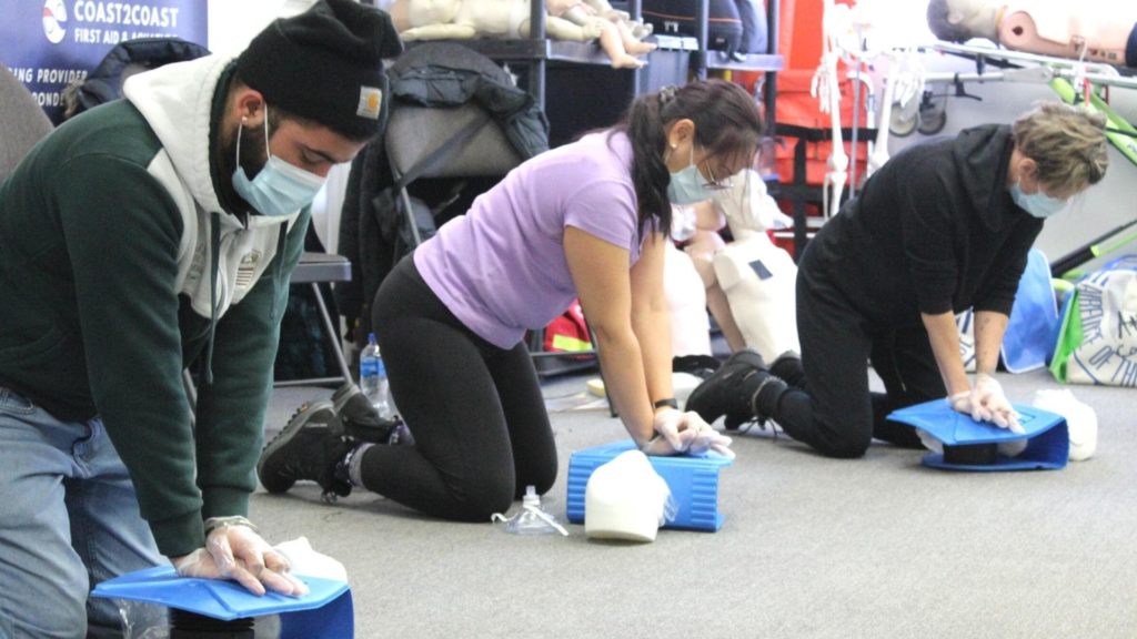 Three people wearing masks and gloves practice CPR on mannequins in a training session. They are kneeling and performing chest compressions. The setting appears to be a classroom or training facility with instructional materials visible.