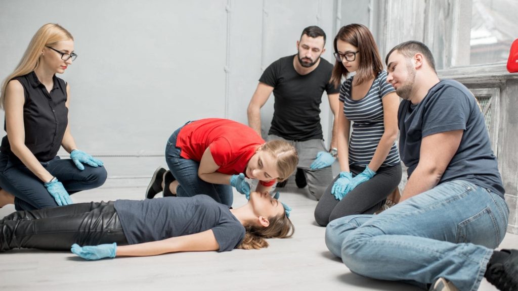Group of people in a first aid class practicing CPR on a person lying on the floor. One individual is administering chest compressions while others observe, all wearing gloves. The setting is a bright room with a large window.