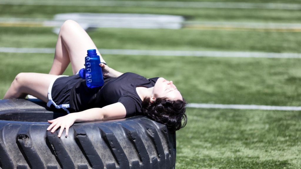 A person in a black shirt and shorts lies on a large tractor tire, holding a blue water bottle. They are on a grassy field under bright sunlight, appearing to rest or relax.