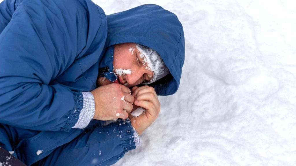 A person wearing a blue jacket is lying on their side in the snow. Their head is covered with the jacket's hood, and their face is partially covered with snow. They appear to be trying to keep warm.