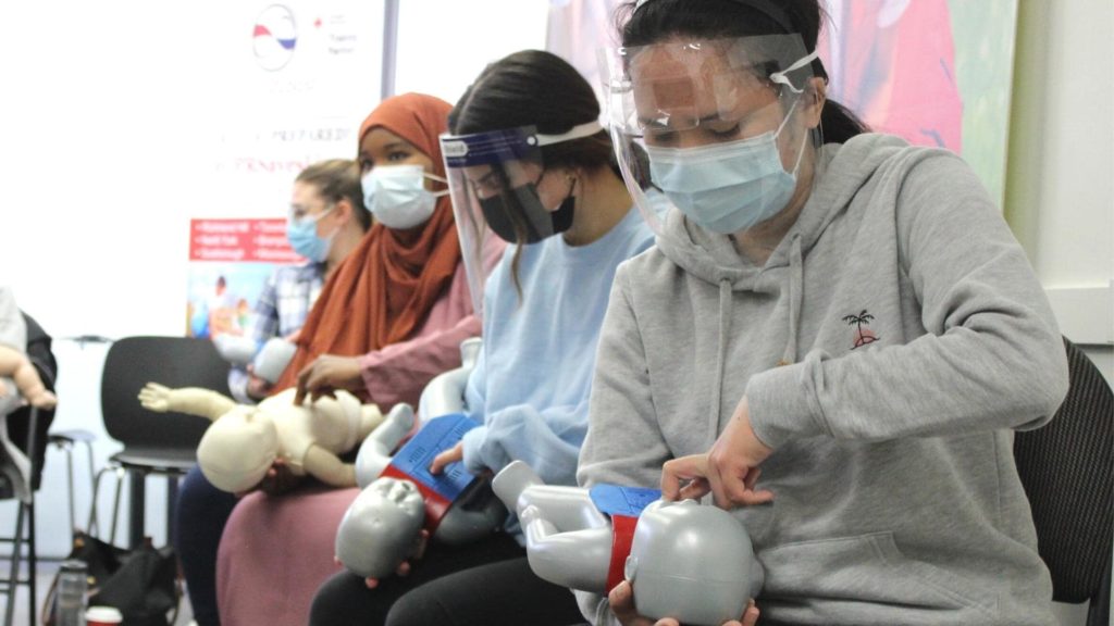 A group of people wearing face masks and shields practice CPR on infant mannequins, seated in a classroom setting. They are focused and engaged in learning the life-saving technique. Posters are visible in the background.