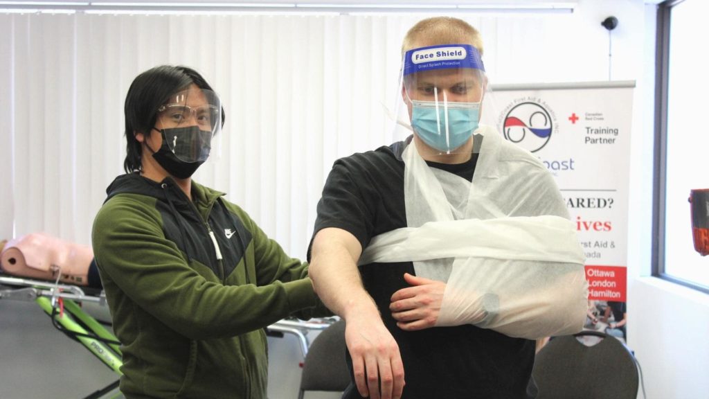 Two people in a training session practice applying a triangular bandage to create an arm sling. One person assists, while the other has their arm wrapped and secured. Both wear face masks and transparent face shields. A first aid poster is visible behind them.