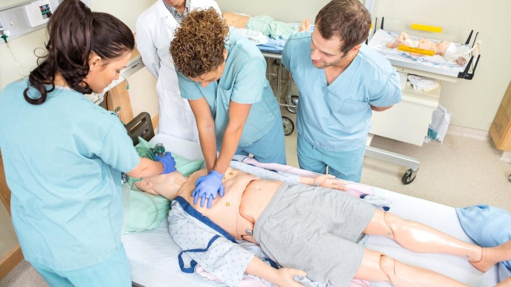 Four medical professionals in scrubs practice CPR on a medical dummy in a clinical setting. One person performs chest compressions while another manages the airway. Two others observe closely, with medical equipment visible in the background.