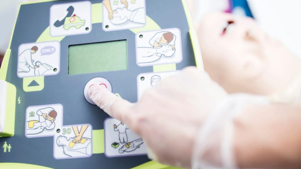 Close-up of a person's gloved hand pressing a button on an automated external defibrillator (AED). The device features illustrated instructions for CPR and defibrillation on its interface.