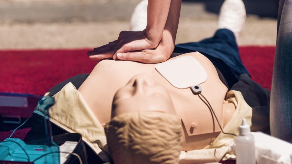 A person is performing CPR compressions on a training mannequin positioned on a red carpet. The mannequin has a defibrillator pad attached to its chest.
