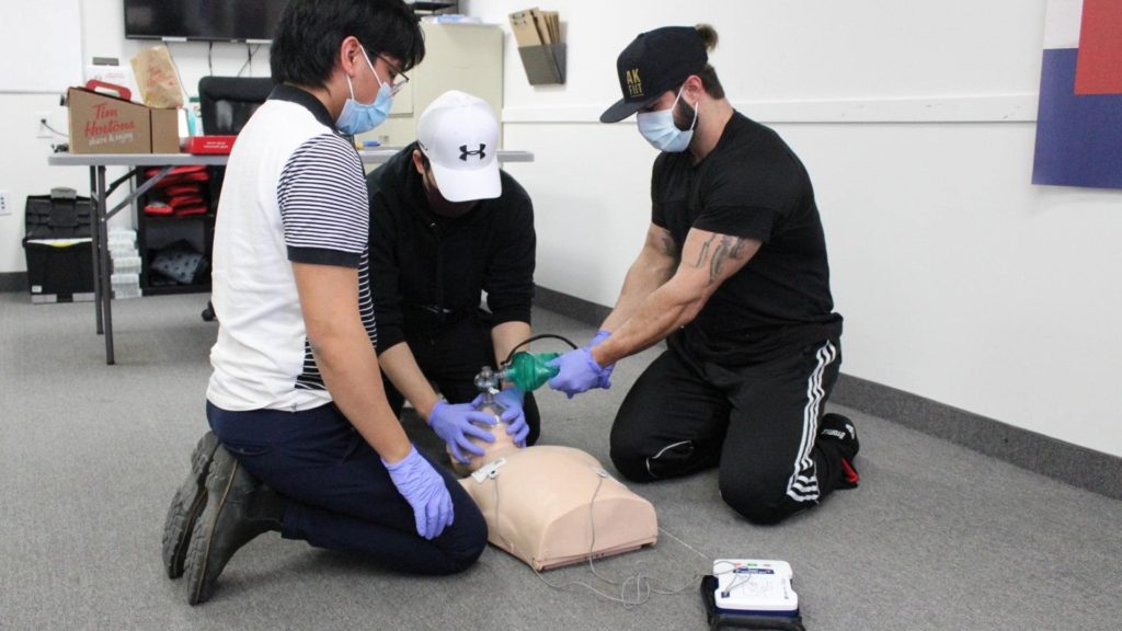 Three individuals wearing masks and gloves kneel on the floor practicing CPR on a mannequin. One person uses a bag valve mask while another operates an AED. They are in a room with a table and supplies in the background.