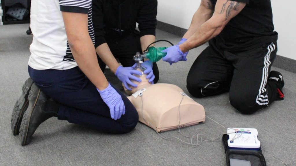 Three individuals in a training session practice CPR on a dummy. One person compresses the chest, another holds a bag valve mask, and a third guides. An automated external defibrillator (AED) is visible on the floor. All wear gloves.