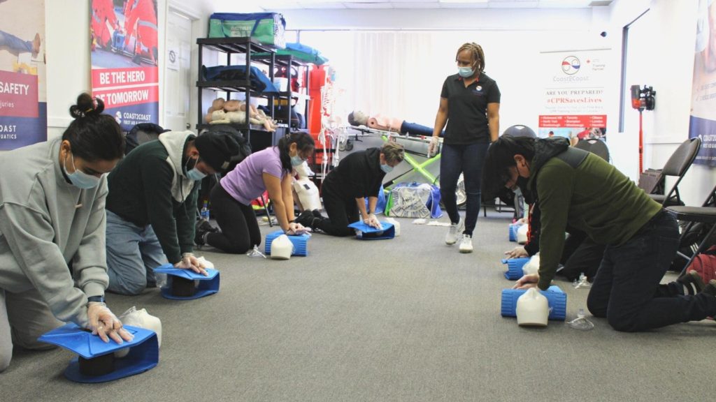 A group of people practicing CPR on mannequins in a classroom setting. They are kneeling on the floor, each with a CPR dummy. An instructor stands, observing and guiding them. Everyone is wearing masks. Educational posters are visible on the walls.