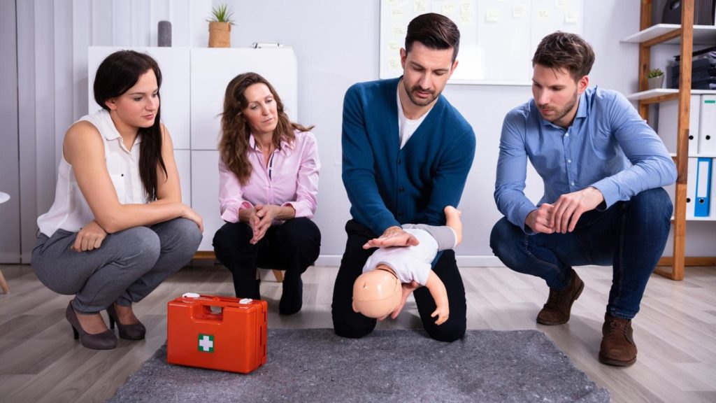 A man demonstrates infant CPR on a doll while three people watch attentively. A first aid kit is on the floor nearby. They are in a bright room with shelves and a whiteboard in the background.