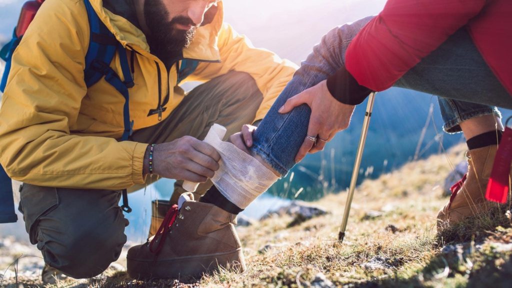 A person in a yellow jacket crouches down, wrapping a bandage around another person's ankle. The injured person sits on a grassy hillside, wearing a red jacket and jeans. Walking poles are nearby, with a sunny outdoor setting in the background.