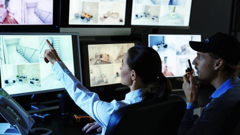 Two security personnel monitor multiple surveillance screens. One points at a screen, while the other speaks into a walkie-talkie. The screens display various indoor and outdoor scenes, suggesting a focus on security operations.