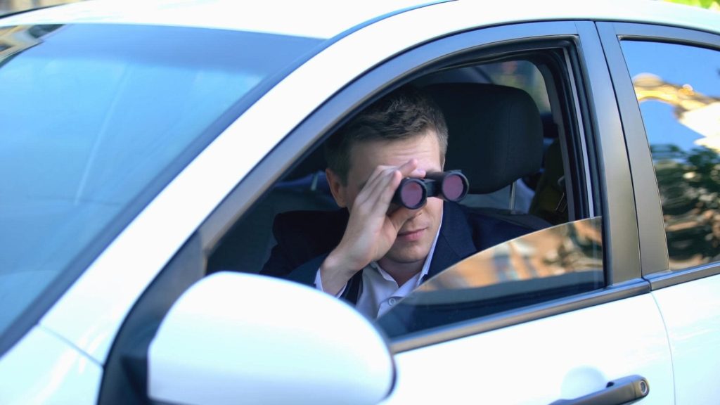 A person sits in the driver's seat of a car, looking through binoculars out the open window. The car is white, and the scene suggests they might be observing someone or something outside.