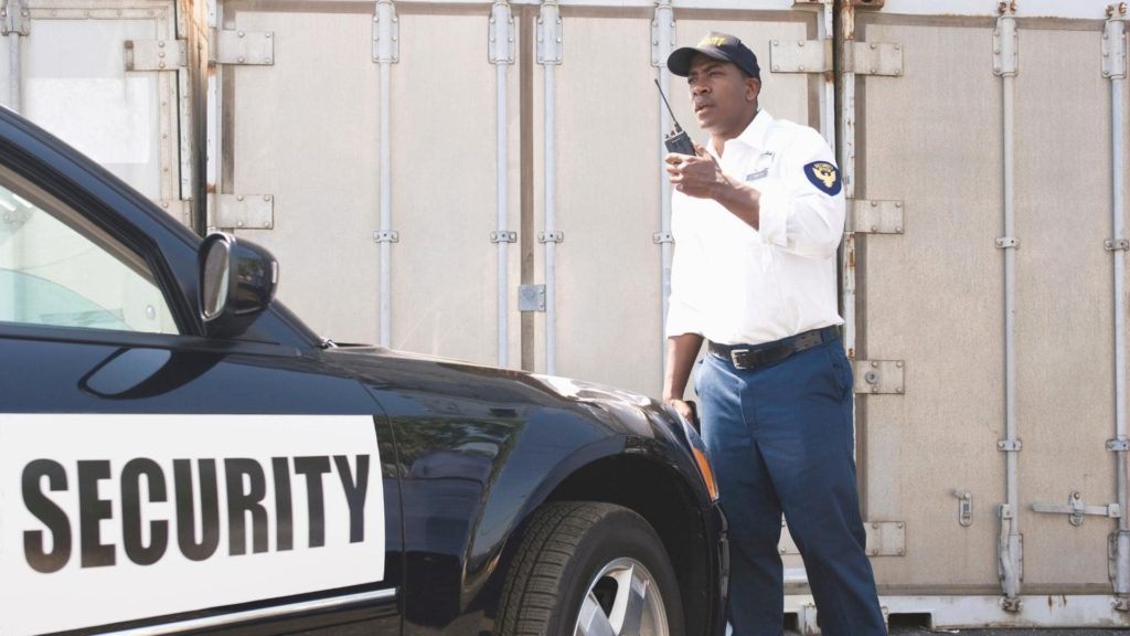 A security guard in a white shirt and cap speaks into a walkie-talkie while standing next to a black and white security vehicle. The background features a large metal industrial door.