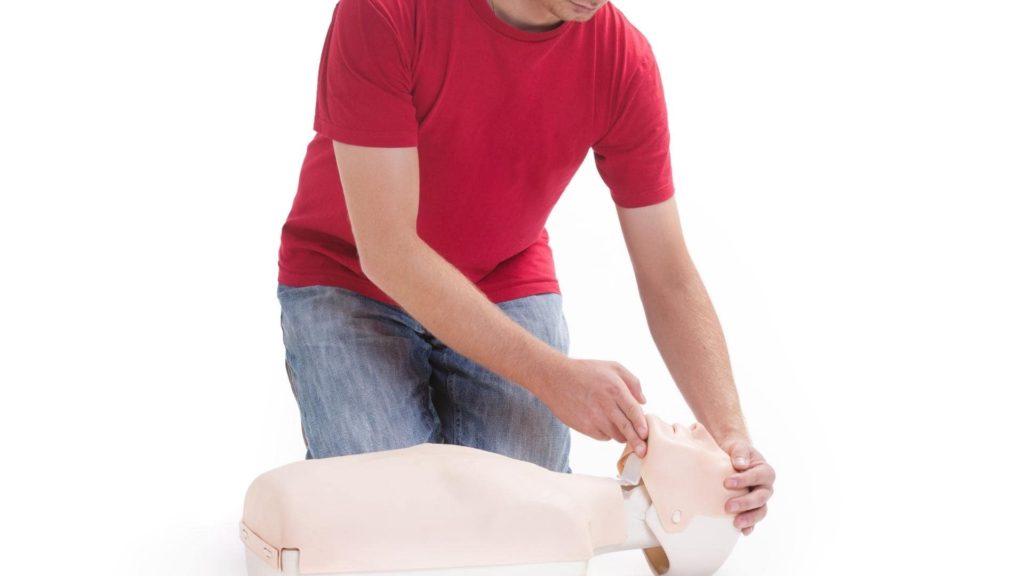 A person in a red shirt and jeans kneels on the floor, practicing CPR on a dummy. They are tilting the dummy's head back, demonstrating a proper airway technique. The background is plain white.