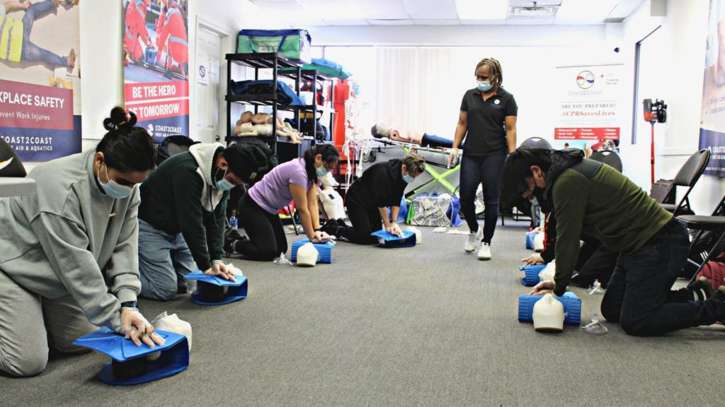 A group of people kneeling on the floor practicing CPR on mannequins in a classroom setting. An instructor stands nearby observing. They are all wearing masks, and safety posters are visible on the walls.