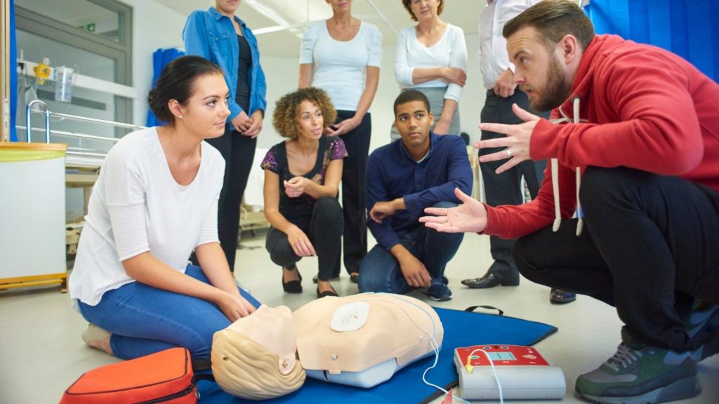A group of people in a classroom setting are gathered around a CPR manikin. A man in a red hoodie is demonstrating CPR techniques, while a woman kneels beside the manikin. Others watch attentively.