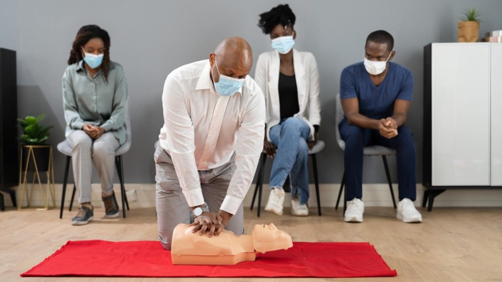 A person in a white shirt demonstrates CPR on a mannequin placed on a red mat, while three seated individuals wearing masks observe attentively. They're in a room with light walls and a cabinet.