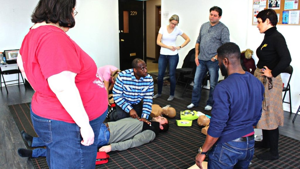 A group of people in a classroom setting are learning CPR techniques. They are gathered around dummies on the floor. One person is kneeling, practicing chest compressions, while others watch and learn.