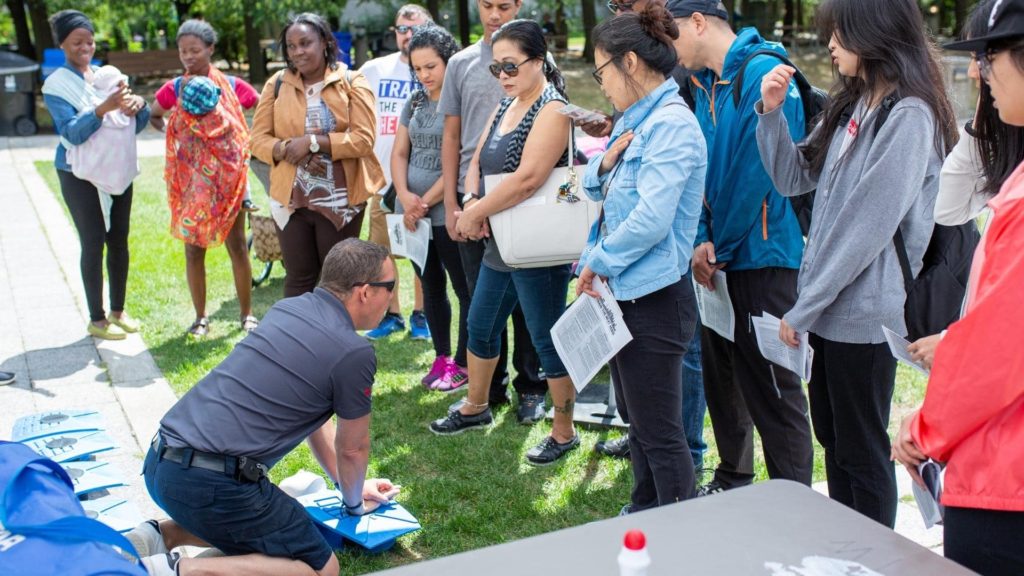A group of people outdoors attentively watch a man kneeling on the grass, demonstrating CPR on a mannequin. The group, holding papers, observes the training session. Sunlight filters through trees in the background.