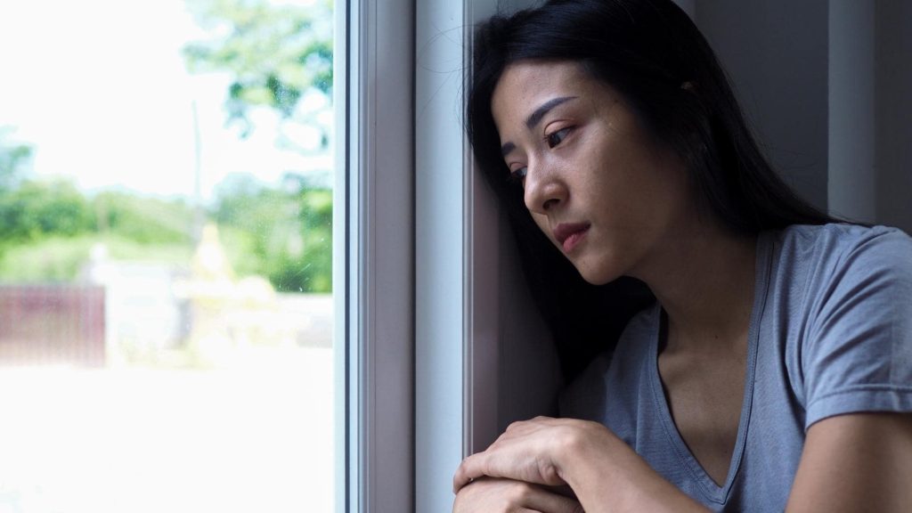 A woman with long dark hair leans against a window, looking outside with a thoughtful expression. She is wearing a gray shirt, and the scene outside is blurred greenery. The lighting is soft, creating a calm atmosphere.