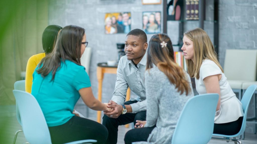 A diverse group of five people sit in a circle, engaged in conversation. They are seated on light blue chairs in a bright room with a gray brick wall and framed photos in the background. A potted plant partially obscures the view.