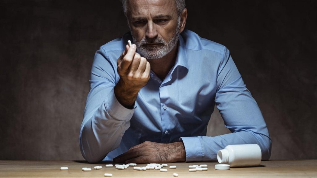 A man with gray hair and a beard wearing a blue shirt sits at a table in a dimly lit room. He holds a pill in his hand, looking at it thoughtfully. Several pills are scattered on the table beside a white bottle.