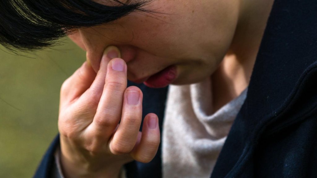A person is holding their face with their hand, fingers pinching the bridge of their nose. They appear to be concentrating or distressed. They have dark hair and are wearing a dark jacket over a light top.