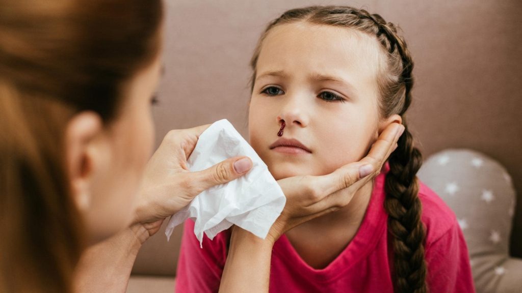 A child with braided hair and a small amount of blood from her nose sits calmly as an adult gently wipes her face with a tissue. She is wearing a pink shirt and appears to be indoors.