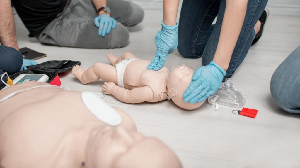 Individuals wearing gloves practice CPR on infant mannequins on a wooden floor. Equipment like a defibrillator and resuscitation mask are nearby.
