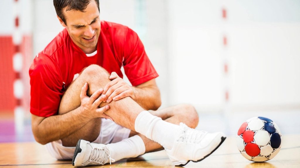 A man in a red shirt and white sneakers sits on an indoor sports court, holding his knee in pain. A colorful soccer ball is nearby. The blurred background suggests a gym or sports hall setting.