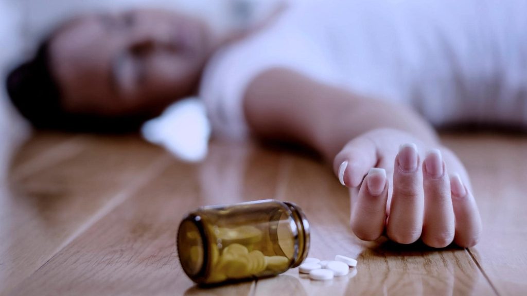 A person lies unconscious on the wooden floor, with an outstretched arm and scattered white pills near an overturned brown bottle. They are wearing a white shirt, and a crumpled tissue is nearby.