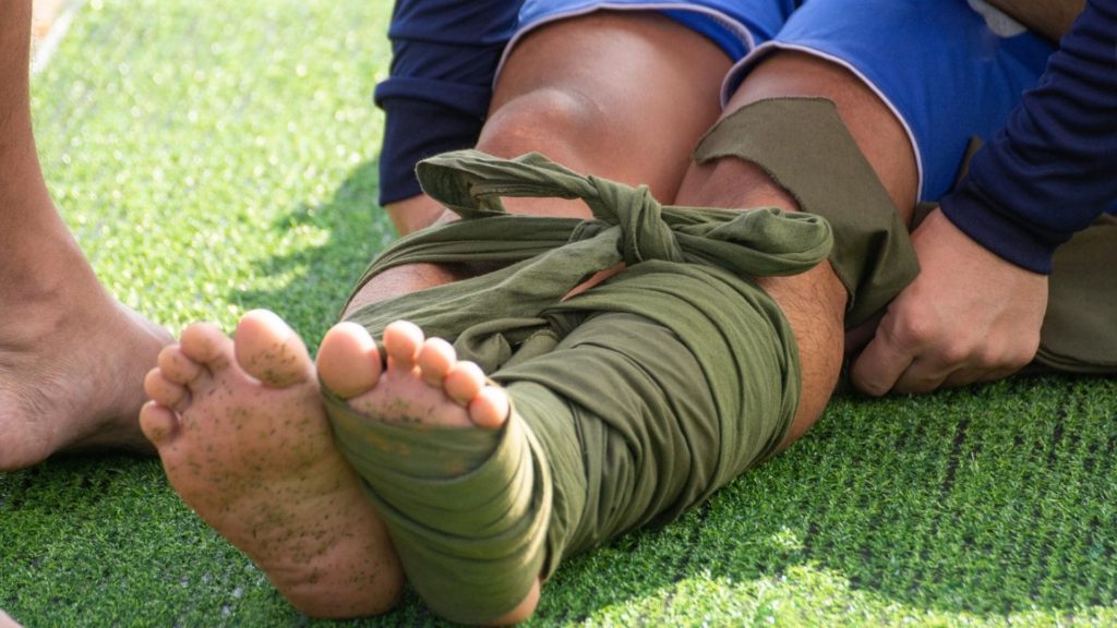 A person sitting on grass with one leg wrapped in green fabric for support or injury. Another person assists in adjusting the fabric. The person is barefoot, with visible green specks on the sole. The individual is wearing blue shorts.