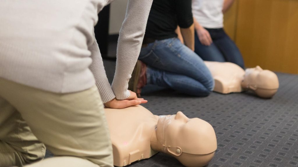 People practicing CPR on mannequins placed on a carpeted floor. The individuals are kneeling and performing chest compressions as part of a training session.