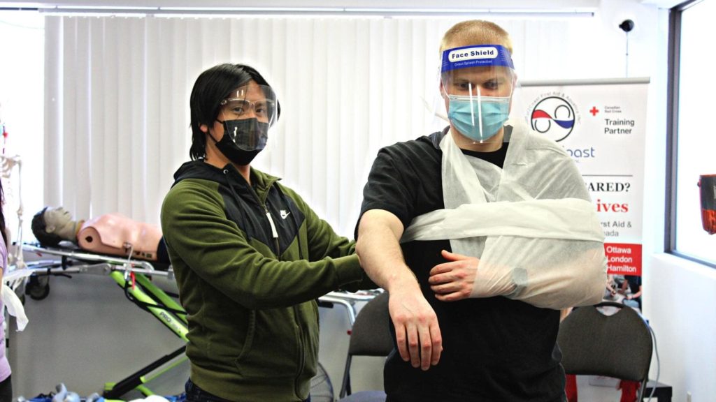 Two people practice a first aid technique. One wears a mask and supports a man with his arm in a sling, also wearing a face shield and mask. A CPR mannequin and a first aid training sign are in the background.