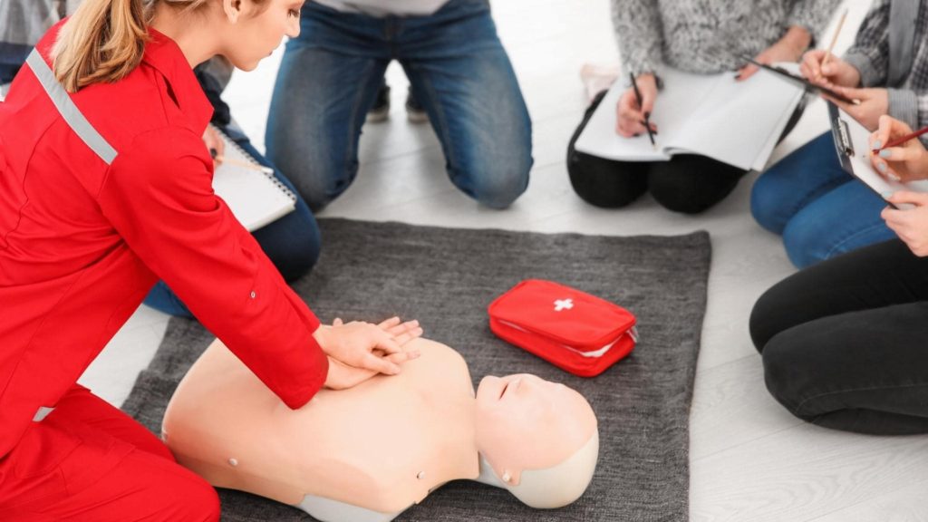 A woman in a red uniform demonstrates CPR on a medical training dummy. She is surrounded by sitting individuals taking notes. A red first aid kit is next to the dummy.