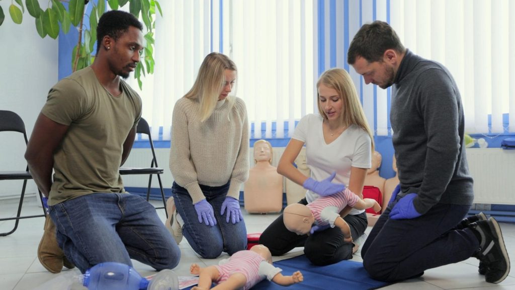 Four people practice infant CPR on mannequins in a room with chairs and a large plant. They wear gloves and are focused on the activity, demonstrating rescue breathing and chest compressions.