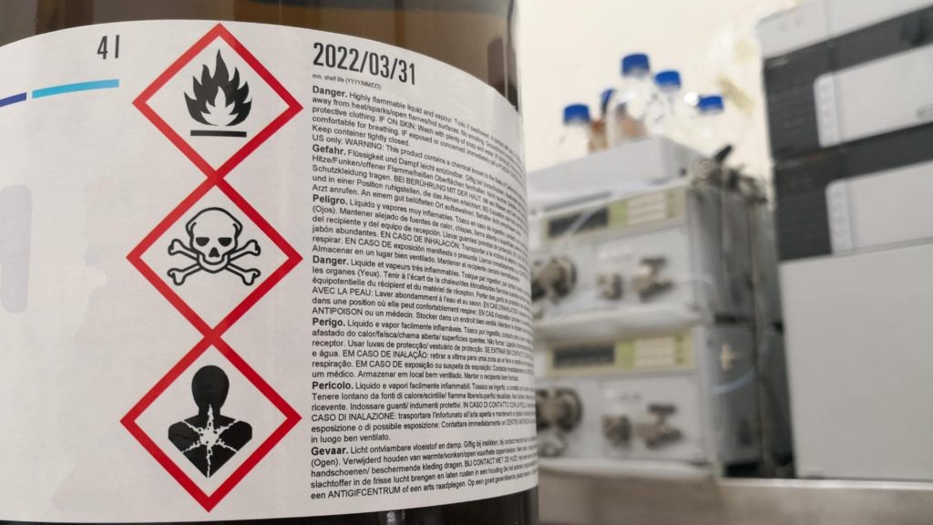 A close-up of a chemical container's label featuring hazard symbols: flame, skull and crossbones, and health hazard. It includes a lot of small text and the date 2022/03/31. Laboratory equipment is blurred in the background.