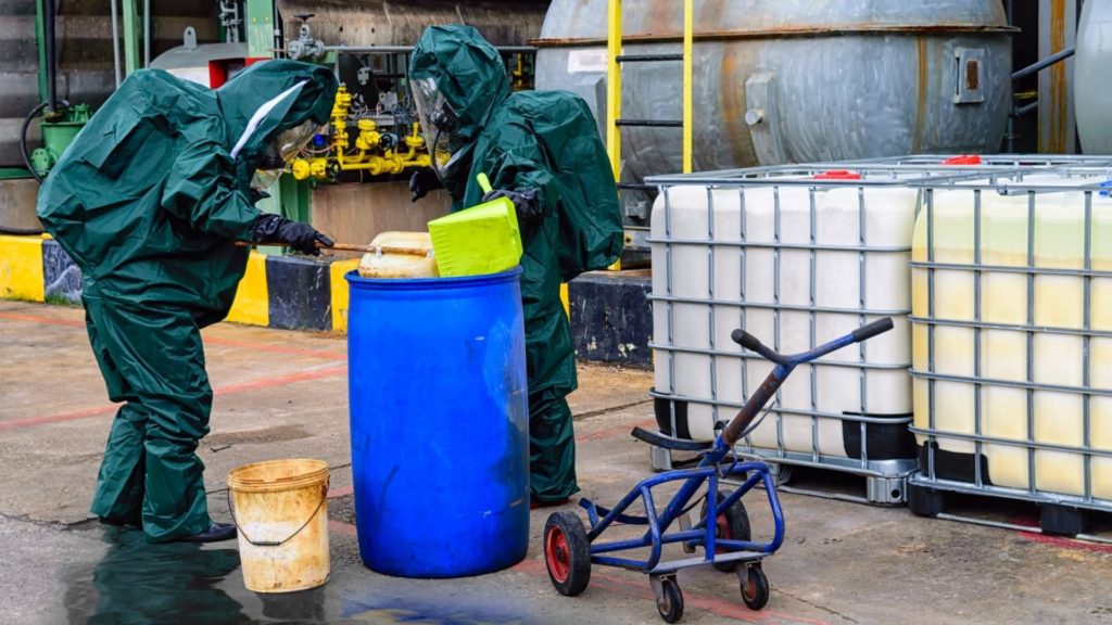 Two people in protective suits handle chemicals, pouring contents from a green container into a large blue barrel. Nearby are large liquid containers and a dolly, suggesting an industrial setting.