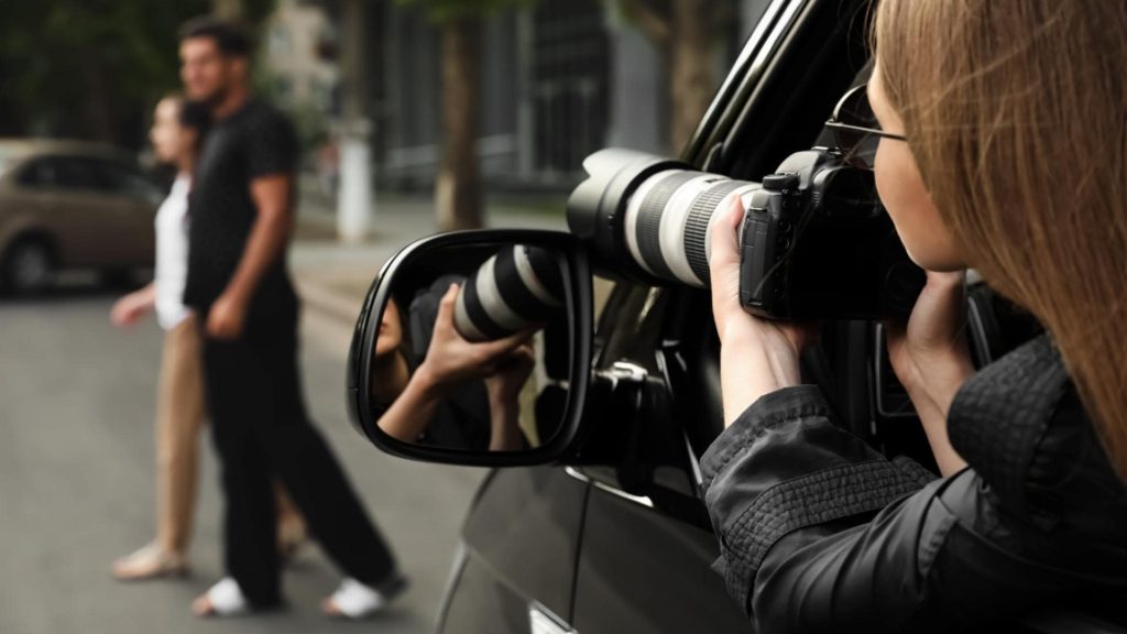 A person with long hair is sitting inside a car, holding a camera with a large lens, focusing on an out-of-focus couple walking on the sidewalk. The scene is captured through the open car window.