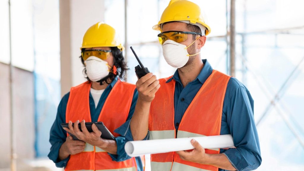 Two construction workers wearing yellow helmets, safety goggles, masks, and orange vests stand on-site. One holds blueprints and a walkie-talkie, while the other uses a tablet. Scaffolding is visible in the background.